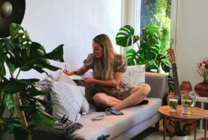 A woman sits on her couch surrounded by plants, writing in a notebook