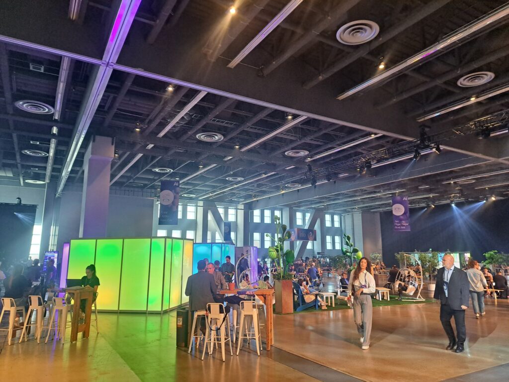 A snapshot of the conference space. High ceilings, and tables and chairs full of people.