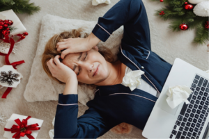 Woman lays on the ground with hands on her head in stress with a laptop on top of her, surrounding by Christmas clutter.
