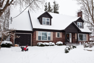 The exterior of a suburban home after a fresh snowfall. A snow blower sits on the lawn.