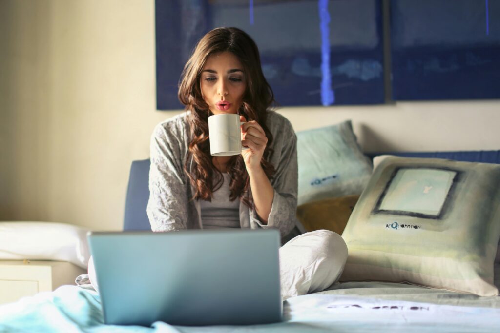 Woman on bed looking at computer screen with cup of coffee in hand