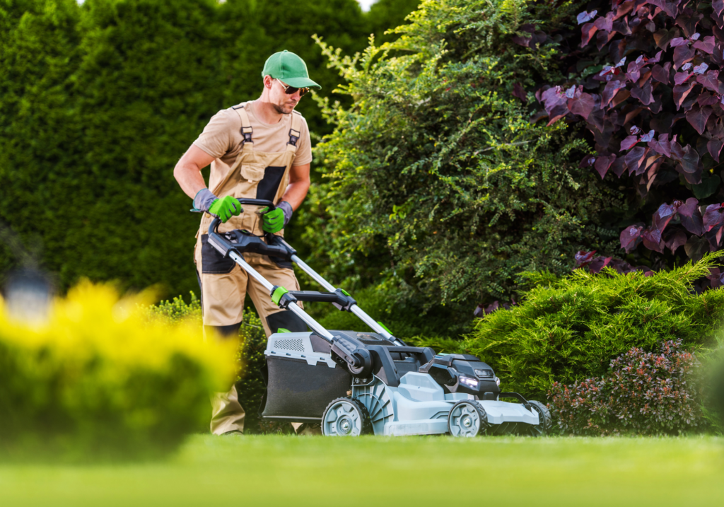 A man in working gear pushes a lawnmower through a lush yard.