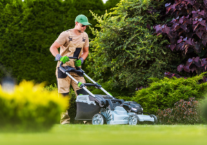 A man in working gear pushes a lawnmower through a lush yard.